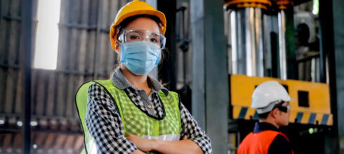 female worker in industrial warehouse wearing safety vest, hard hat, glasses and COVID mask