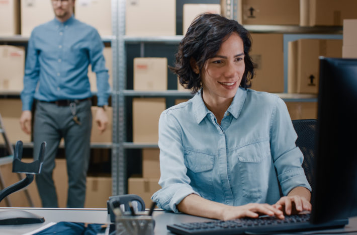 Female Inventory Manager Sitting at Her Desk and Using Personal Computer, Worker Puts Packages on the Designated Shelf. In the Background Rows of Cardboard Boxes with Products Ready For Shipment.