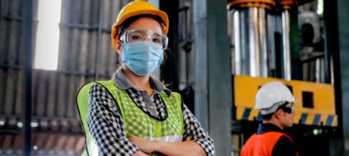 female worker in industrial warehouse wearing safety vest, hard hat, glasses and COVID mask