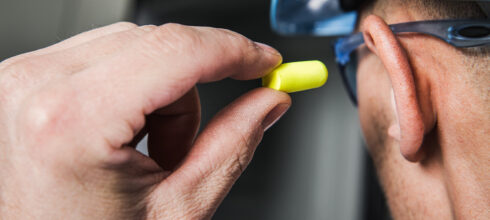 A person holding a yellow earplug near their ear, preparing to insert it, with protective eyewear and a hard hat visible in the background.