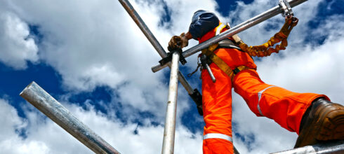 A worker in orange safety gear and a harness climbing on scaffolding against a blue sky with clouds, illustrating the importance of securing tools while working at height.