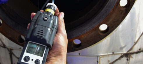 A close-up image of a handheld gas detection device being held near industrial equipment, displaying readings on its digital screen.