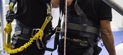 Two workers wearing safety harnesses with visible D-rings and lanyards, illustrating proper fall protection equipment use at height.