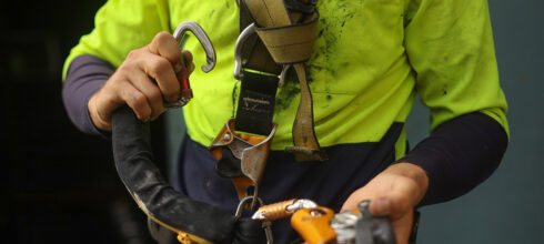 A worker in high-visibility clothing inspecting a fall protection harness and carabiner, emphasizing proper safety equipment use.