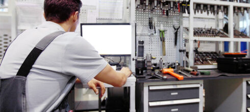 Worker in a tool crib reviewing setup on computer with tools and equipment organized on workbench – re-evaluating tool crib setup.