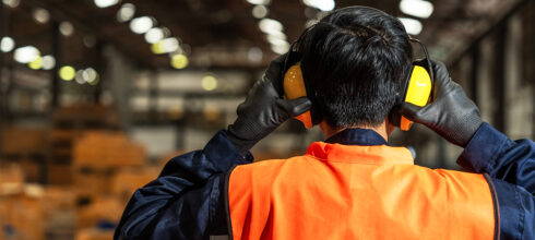“Warehouse worker wearing high-visibility vest and protective earmuffs inside an industrial facility.