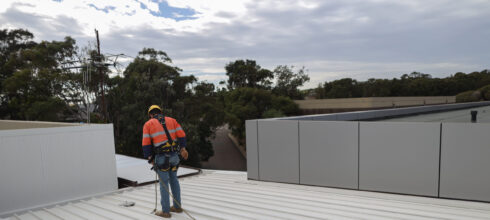 Worker wearing safety gear and a harness attached to anchor points while inspecting or working on a metal rooftop.