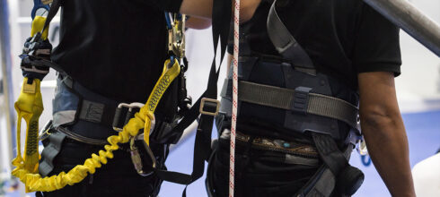 Two workers wearing safety harnesses with visible D-rings and lanyards, illustrating proper fall protection equipment use at height.