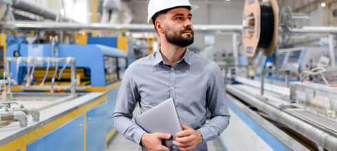 Manufacturing supervisor holding a laptop while inspecting production equipment for potential lean manufacturing inefficiencies.
