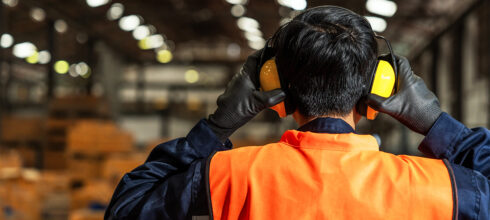 “Warehouse worker wearing high-visibility vest and protective earmuffs inside an industrial facility.